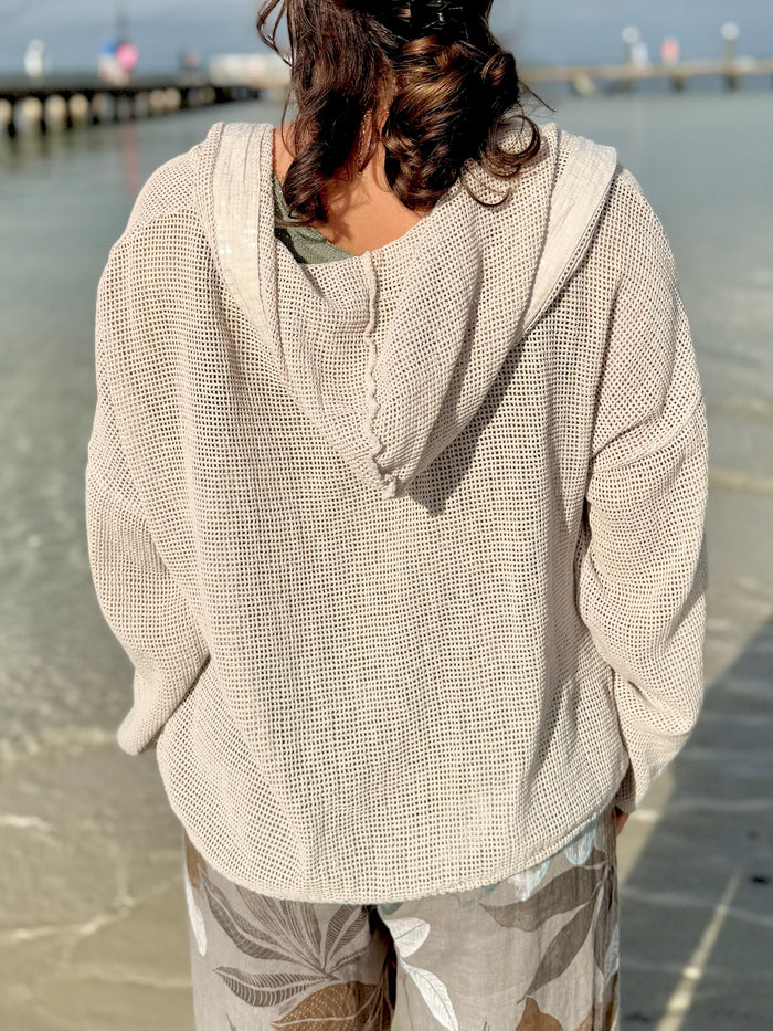 Back view of the Gia Mesh Hooded Jacket in beige, showing the loose open-knit texture and relaxed hood as the model stands by the water in sunlight.