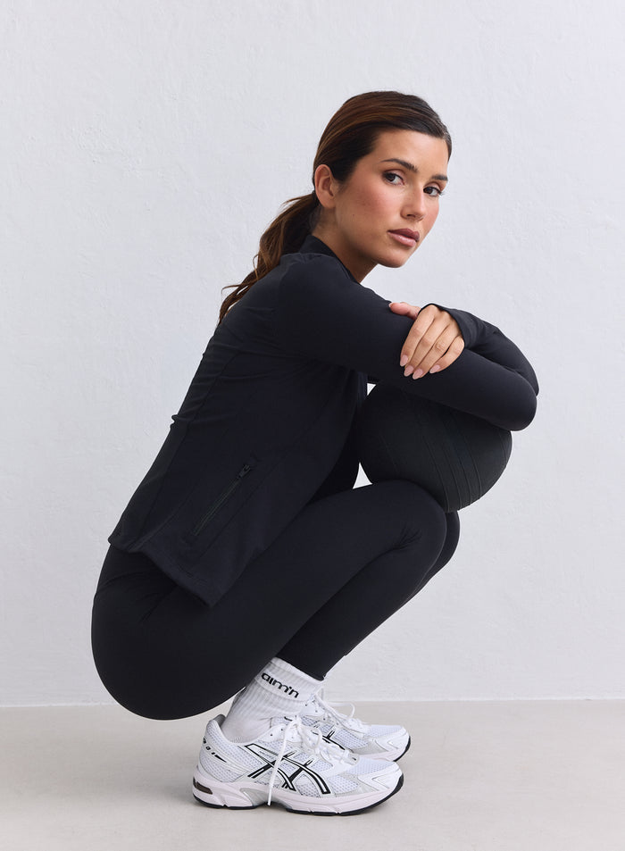Nainen mustassa aim'n Sense Zip Jacketissa ja second skin -tuntuisissa trikoissa kyykistyneenä pallon kanssa. [XS-165cm]